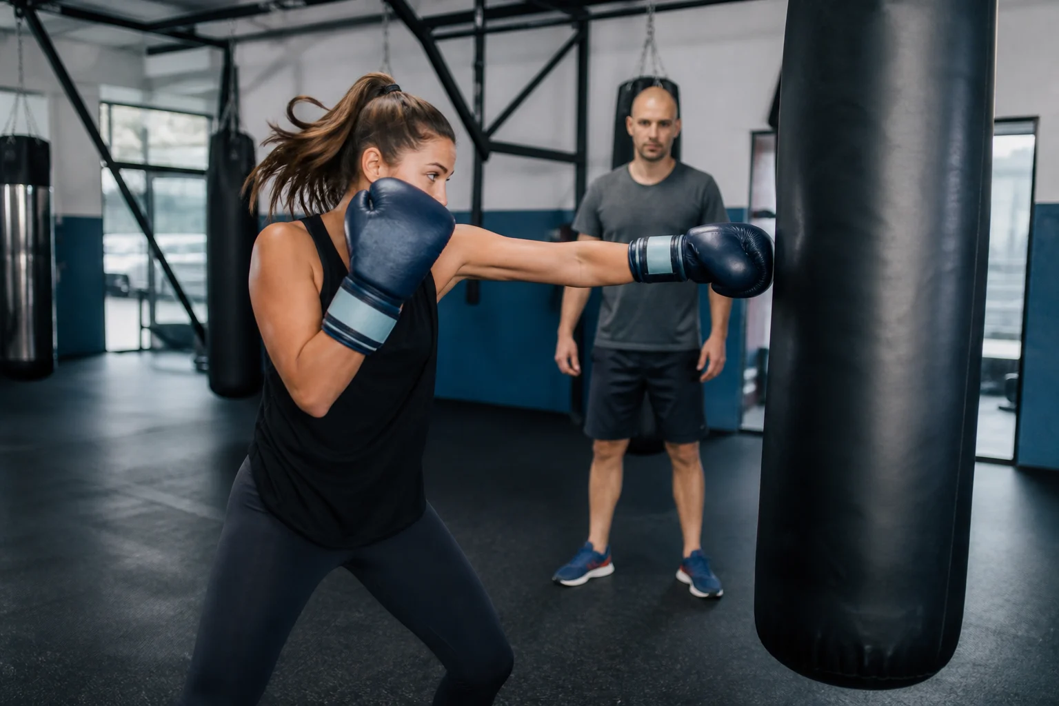 Mujer golpea una bolsa de boxeo con guantes mientras un profesor supervisa